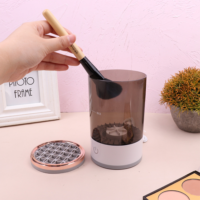 Hand holding a brush over a small cylindrical device with a transparent top on a light pink surface.